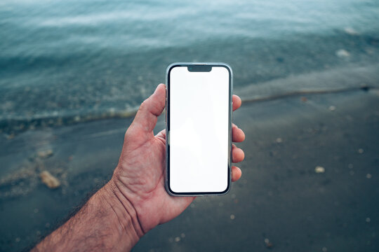 POV shot of a person holding a smartphone with a blank white screen mockup on a beach next to calm sea waters at dawn.