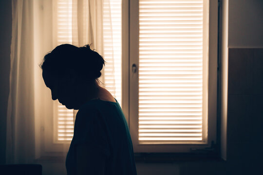 Silhouette of a woman standing in profile indoors in front of a window with horizontal blinds.