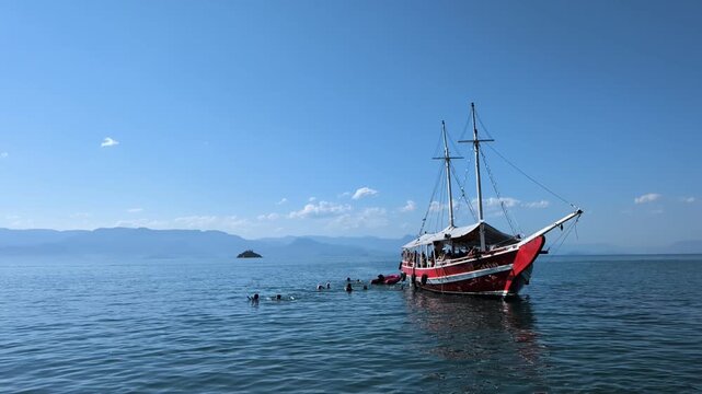 Handheld slow motion of a traditional red schooner boat floating on calm blue waters in Paraty, Rio de Janeiro, with tourists swimming next to it on a clear sunny day with mountains behind.