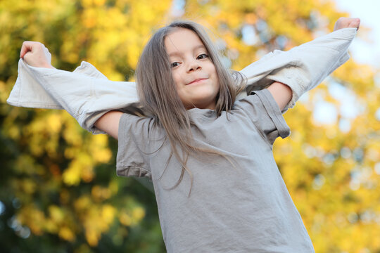 Portrait of a happy girl raising her arms above her head and looking at the camera outdoors with colorful autumn trees in the background, expressing joy, freedom and childhood happiness