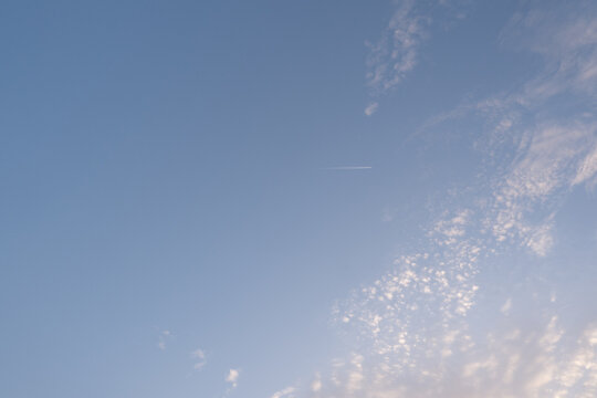 A clear blue sky with soft white cirrocumulus clouds and a distant airplane leaving a straight condensation trail, captured during a bright day.