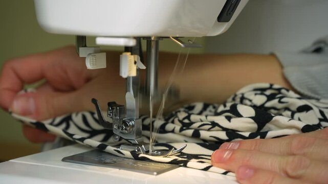 Close-up of hands guiding black and white patterned fabric through a sewing machine during the stitching process. Detailed view of the needle, presser foot, and textile as a garment is being sewn