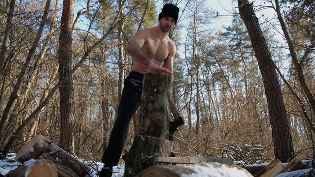 Muscular guy placing a log on a stump in a winter forest. Shirtless young man prepares to chop firewood with axe in snowy woodland. Concept of energy and harmony with nature during cold season