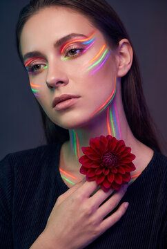 Young woman with creative neon light makeup holding red flower in studio portrait. Concept of rainbow makeup, beauty portrait.