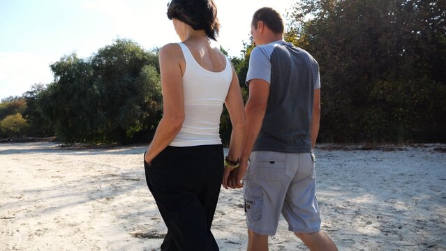 Happy pair going along coast at sunny day. Romantic couple holding hands of each other and walking on sandy beach. Lovers spending time together at resort. Concept of summer vacation or holiday