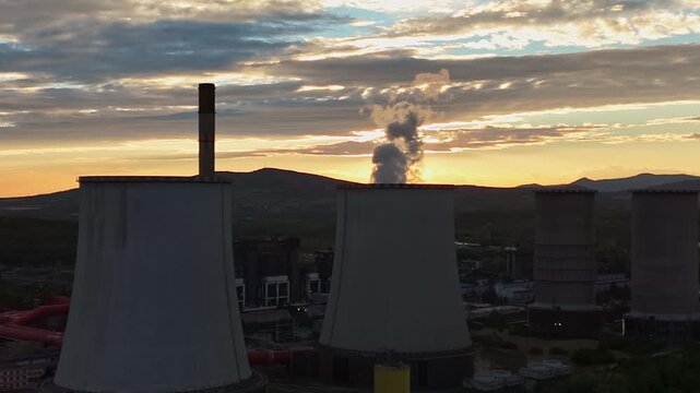 Aerial view of cooling towers and a smokestack at a lignite-fired power plant in Visonta, Hungary, releasing steam over an industrial complex and rural hills at sunset.