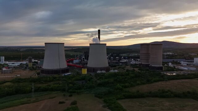 Aerial view of cooling towers and a chimney releasing steam at a lignite-fired power plant near Visonta, Hungary, surrounded by farmland and hills at sunset.