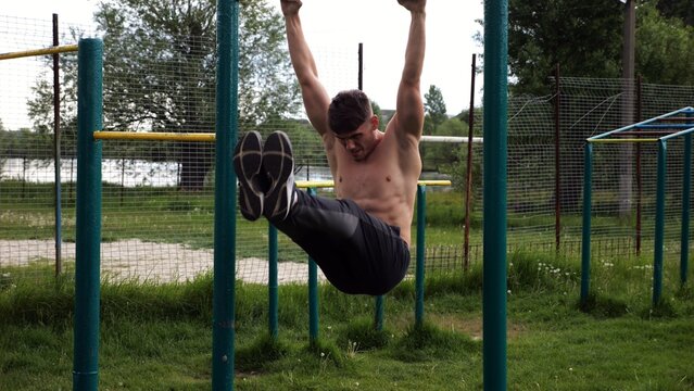 Strong guy training abdominal muscles on horizontal bar at sports ground. Young sportsman exercising on pull-up bar outdoor. Sporty athlete working out at nature. Concept of sportive lifestyle