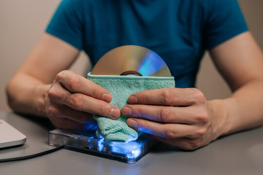 Close-up hand of person wiping a cd or dvd with a soft microfiber cloth, maintaining data storage and playback quality for an external disc drive and laptop setup.