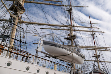 Low angle view of a historic sailing ship deck featuring a white lifeboat on davits near complex mast rigging ropes © MarijaBazarova