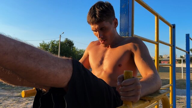 Young shirtless sportsman trains abdominal muscles raising legs on parallel bars at sports ground. Strong man exercising at nature. Sporty athlete working out at nature. Sportive lifestyle