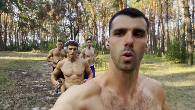 Group of young muscular athletes running at forest path. Active strong men jogging at sunny woodland. Male shirtless sportsmen training at summer nature. Concept of active and healthy lifestyle. POV