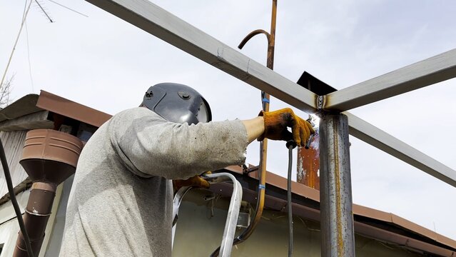Manual laborer in protective mask weld iron detail on carport frame. Professional young repairman with welding machine working at construction. Male welder operating with metal at the site. Close up