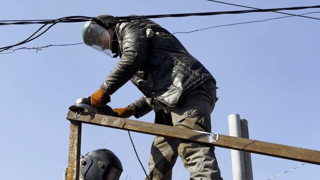 Young mechanic in protective gloves polishing iron using electric grinder wheel at construction. Professional repairman grinding metal details with circular saw on house at sunny day. Slow mo