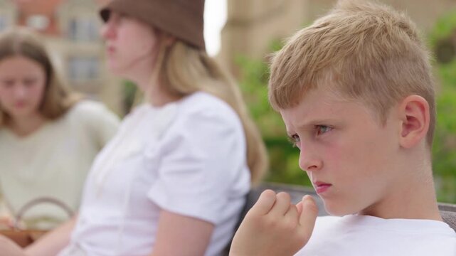 A boy is sitting on a bench next to his mother and sister a family is spending time in the park