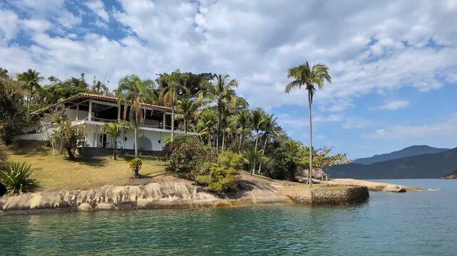 Handheld slow motion from a boat passing by a luxury white beachfront house surrounded by palm trees on Ilha dos Cocos, Paraty, Rio de Janeiro, on a sunny summer day.