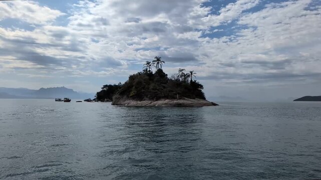 Handheld slow motion from a boat moving right showing a small tropical island with palm trees in Paraty, Rio de Janeiro, with tourist schooners anchored nearby under a cloudy sky.