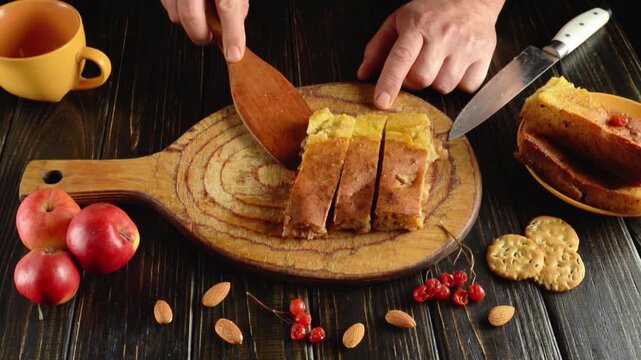 Top view of hands serving sliced fruit bread with a wooden spatula beside a knife, tea, apples, almonds, berries, and crackers on a rustic table