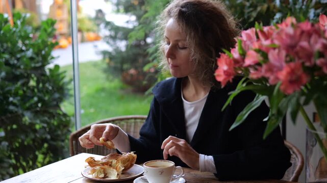 Woman eats pastry at cafe table, pausing to savor her snack with contented, relaxed expression. Solo diner enjoys coffee among green plants and pink blossoms during peaceful leisure break.