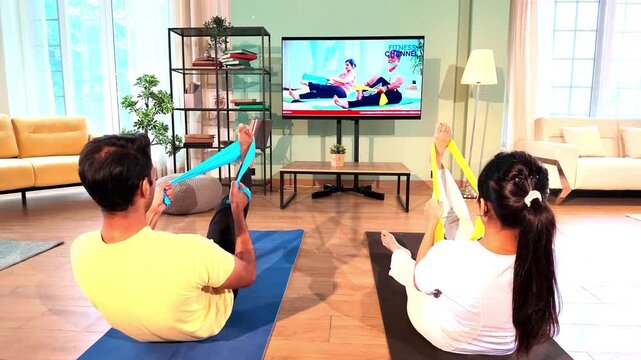 Young Indian couple sitting on the floor and stretching their legs with resistance bands held in both hands while following a TV fitness tutorial in a lavish modern home.