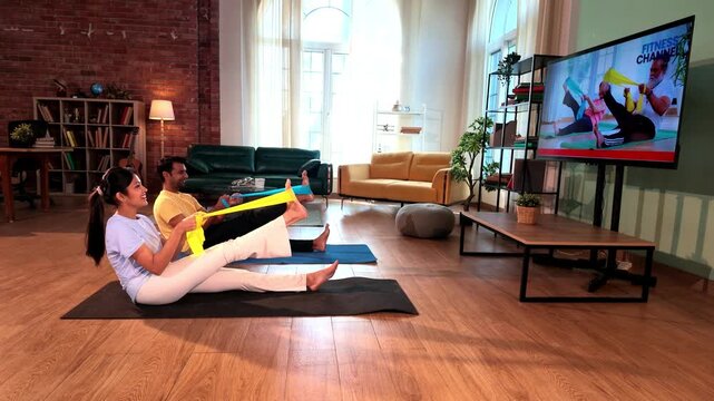 Young Indian couple sitting on the floor and stretching their legs with resistance bands held in both hands while following a TV fitness tutorial in a lavish modern home.