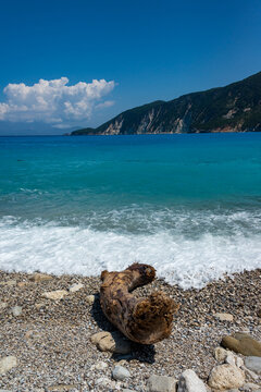 Blue sea and white surf on a pebble beach with a steep wooded coast behind. Taken in Greece