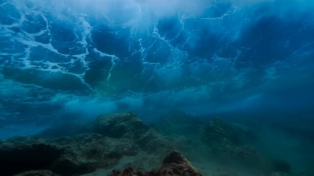 Underwater ocean waves roll over a rocky seabed beneath a foamy sea surface. Nature documentaries, marine conservation campaigns, and oceanography education.