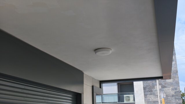 Light gray concrete ceiling of a balcony with round ceiling light, dark gray wall with roller shutter over the apartment balcony door, stone-clad building with glass balcony and air conditioner behind