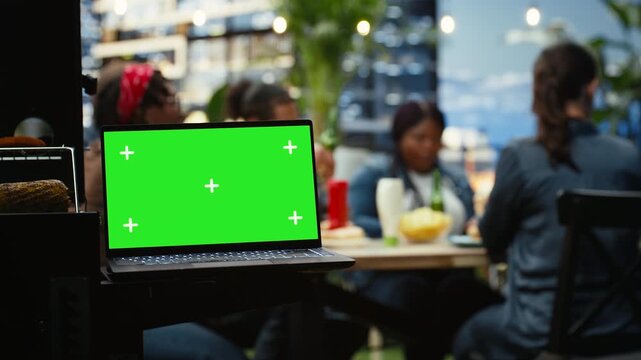 Green screen laptop with chroma key display sits at a rooftop barbecue party, providing editable screen replacement space while friends socialize around food and drinks in an outdoor gathering.