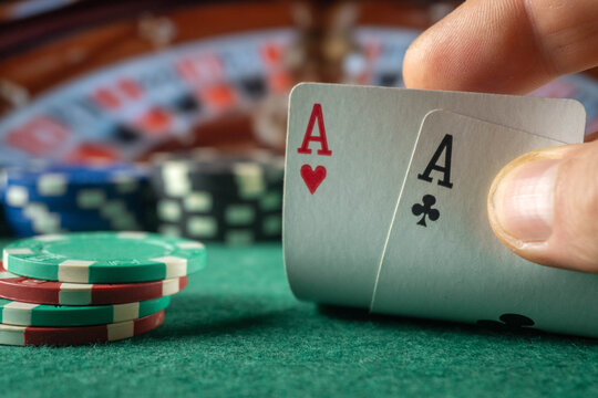 Poker player holds ace of hearts and ace of clubs above felt table, with chips stacked nearby. Pair of aces creates strong starting hand, while shallow focus keeps casino game details softly blurred.