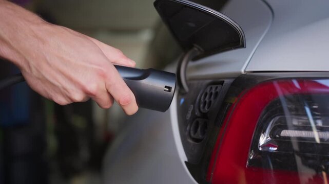 Hand unplugs charging cable from electric car in home garage as charge port flap closes. Selective focus and soft indoor lighting accentuate the modern vehicle tail light.