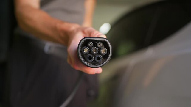 Person holds electric vehicle charging plug in underground garage near car. Shallow depth of field emphasizes the connector pins under soft ambient indoor light.