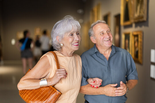 Interested elderly tourists admiring art collection with museum pictures displayed in background
