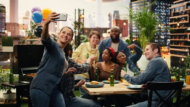 Woman takes a group selfie on her smartphone as smiling friends lean together at a rooftop barbecue party, celebrating friendship and shared fun outdoors.