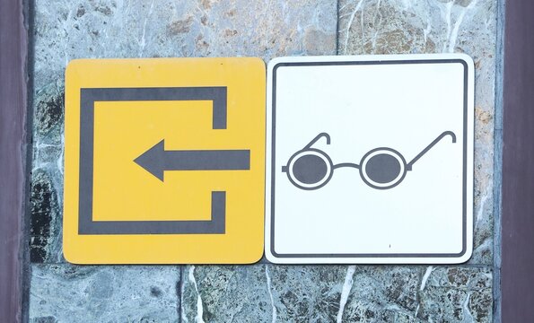 Directional sign with left arrow beside eyeglasses symbol mounted on stone wall. Wayfinding and navigation. Public information signage. Accessibility and visual guidance
