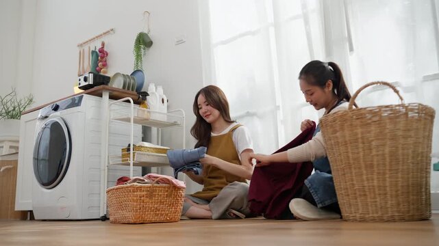 Two young Asian women sit together in laundry room, smiling as they sort and pass folded clothes beside a washing machine and wicker baskets.