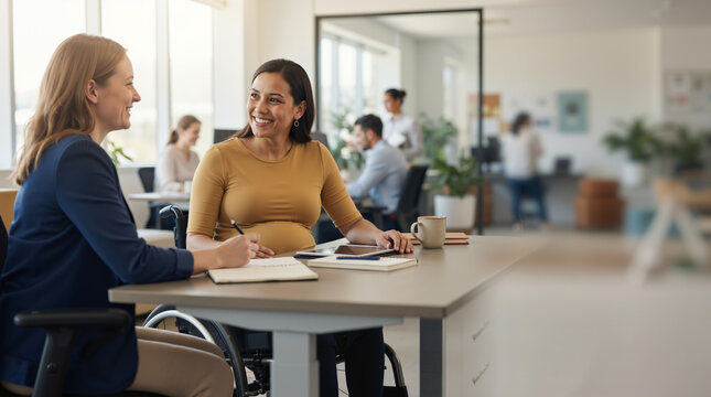 Pregnant businesswoman in a wheelchair talking with a colleague at a desk. Inclusive modern office environment. Diverse corporate teamwork and accessibility concept