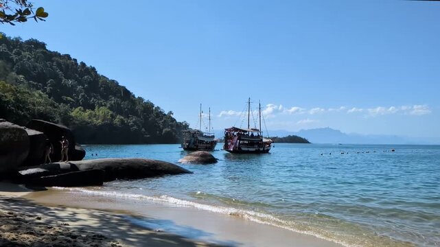 Handheld slow motion from the shore of Praia da Concei&ccedil;&atilde;o in Paraty, Rio de Janeiro, showing two tourist schooner boats anchored in the calm blue sea next to a forested tropical hill on a sunny day.