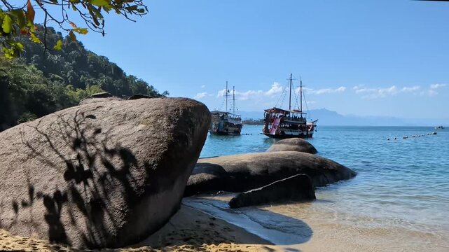 Handheld slow motion from the sandy shore of Praia da Concei&ccedil;&atilde;o in Paraty, Rio de Janeiro, showcasing large shoreline boulders with two tourist schooners anchored in the blue sea behind.