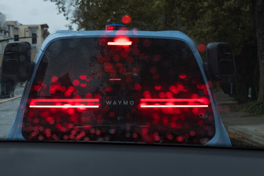 Red brake lights of a Waymo Ojai robotaxi glowing through a rain-covered windshield, raindrops turned into red bokeh circles