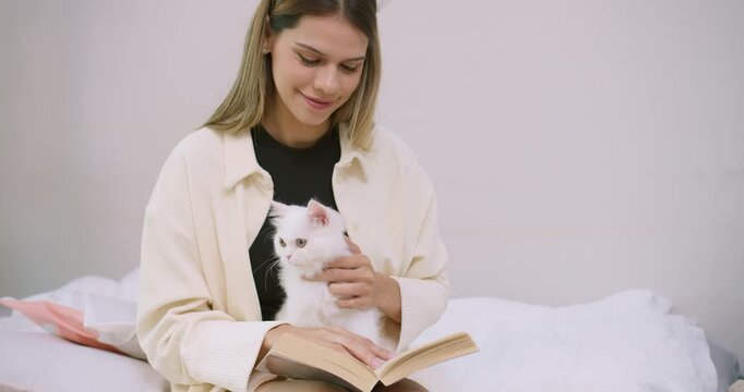 A loving young woman gently pets her adorable white kitten while reading a book in bed, enjoying a peaceful and cozy moment of companionship and leisure at home.