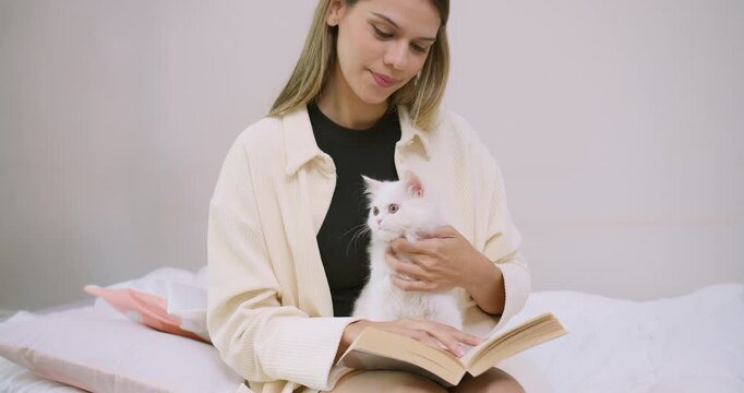 A loving young woman gently pets her adorable white kitten while reading a book in bed, enjoying a peaceful and cozy moment of companionship and leisure at home.