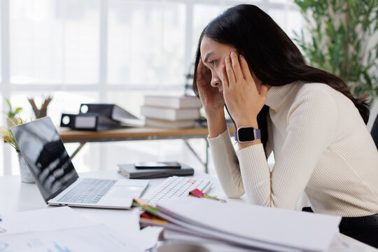 Overworked young asian businesswoman experiencing stress and headache, feeling tired and frustrated at workplace, suffering from burnout while working on laptop in modern office