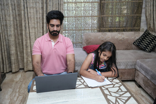 Indian father doing office work on laptop while her little daughter doing school home work. Concept of father and daughter working from home