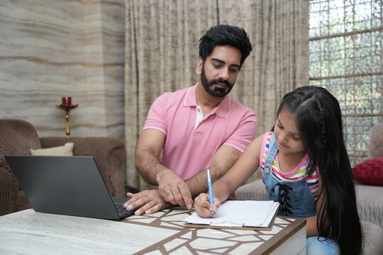 Indian father doing office work on laptop while her little daughter doing school home work. Concept of father and daughter working from home