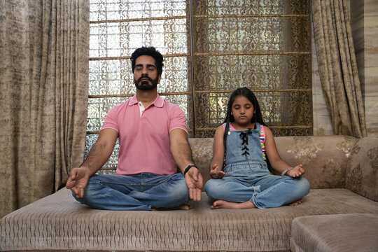 Indian father and her daughter to meditate at home, sitting on couch with closed eyes, keeping padmasana, zen hands. Father and pretty daughter kid doing yoga together. mum teaching child to meditate