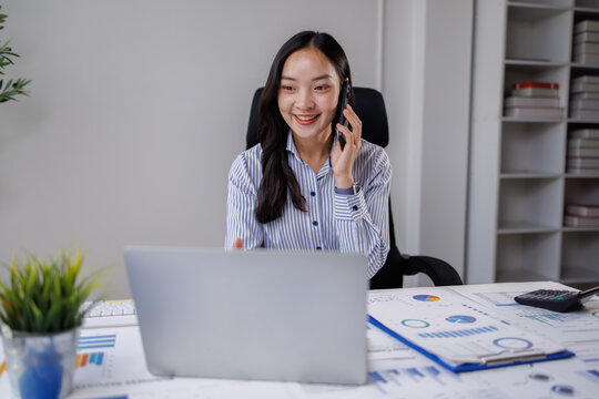 Asian businesswoman smiling during a phone call while working on her laptop and reviewing financial charts and reports at a modern office desk, multitasking confidently