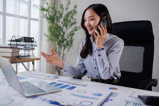 Asian businesswoman talking on a smartphone, discussing data and financial graphs spread across her desk, working efficiently in a bright modern office environment
