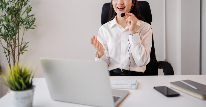Young asian woman wearing a headset and smiling, actively engaged in a call, offering customer service and support from her office desk with a laptop and various work tools