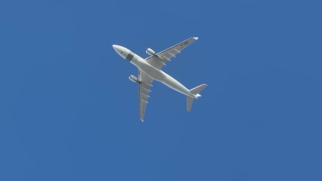 Low angle view of the underbelly of a large commercial passenger jet flying directly overhead against a clear blue sky, showing its wings and engines. 15502  